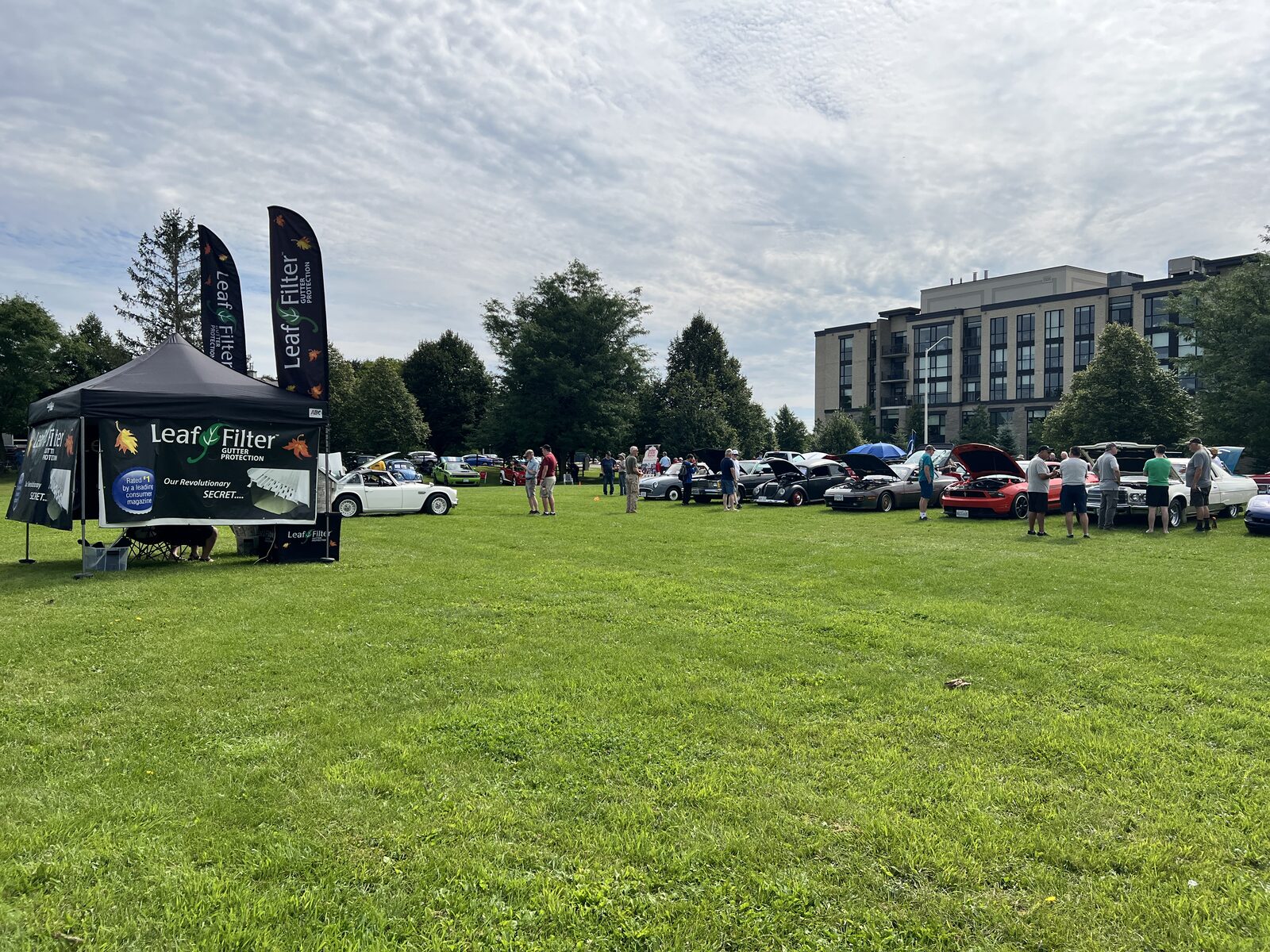 Wide view of the show field at Clarke Fields, a row of cars with their hoods up along the tree line and a sponsor tent at left.