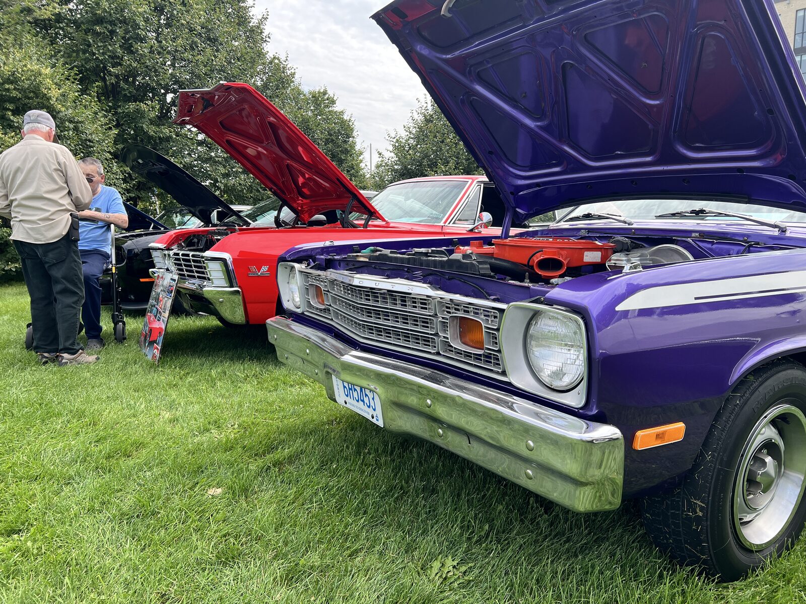 A purple 1970s Plymouth with its hood raised, parked beside a red car with its hood up, both on grass at Clarke Fields.