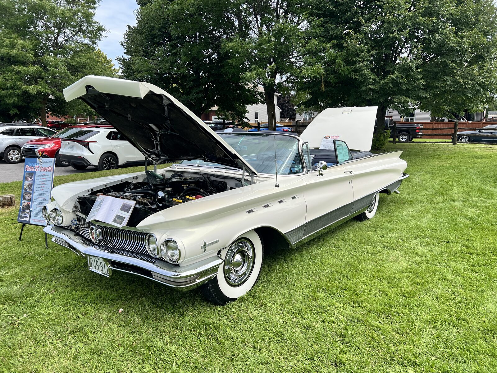 A white 1960 Buick Electra 225 convertible on grass with the hood and trunk both open, its information board standing at the front bumper.