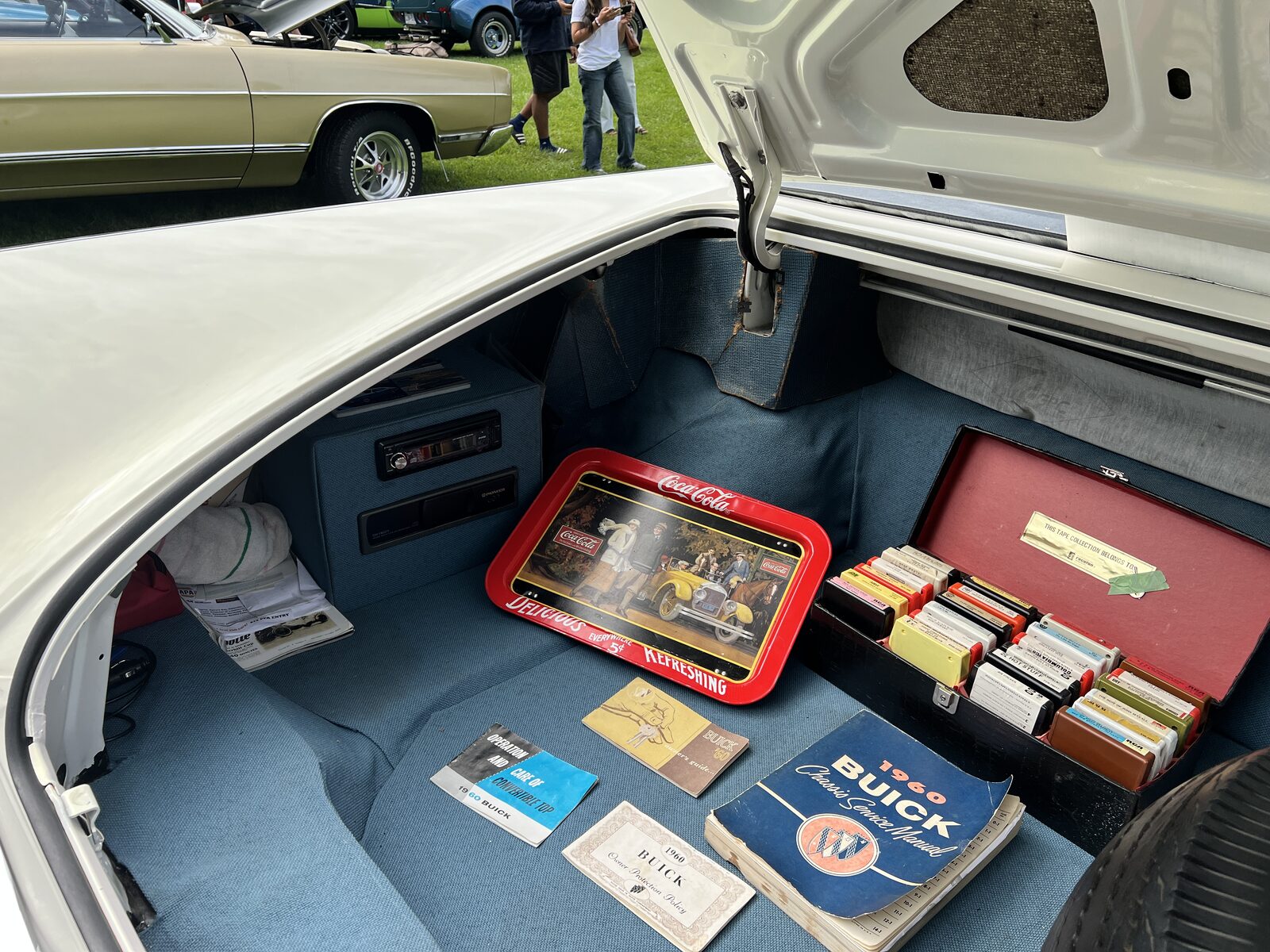 The open trunk of the Buick, laid out with a rack of 8-track tapes, a Coca-Cola serving tray, a 1960 Buick chassis service manual and the original owner booklets.
