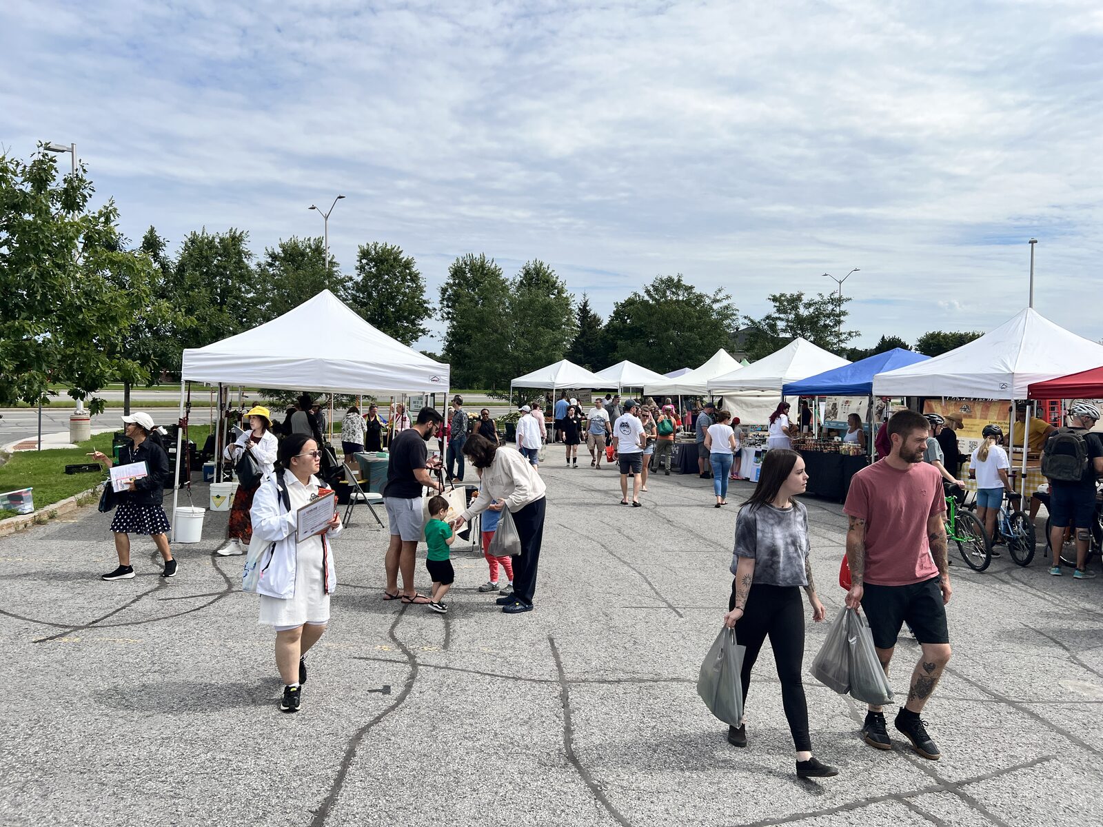 Shoppers crossing the lot between the tents at the Barrhaven farmers' market, with a row of white vendor canopies behind them.