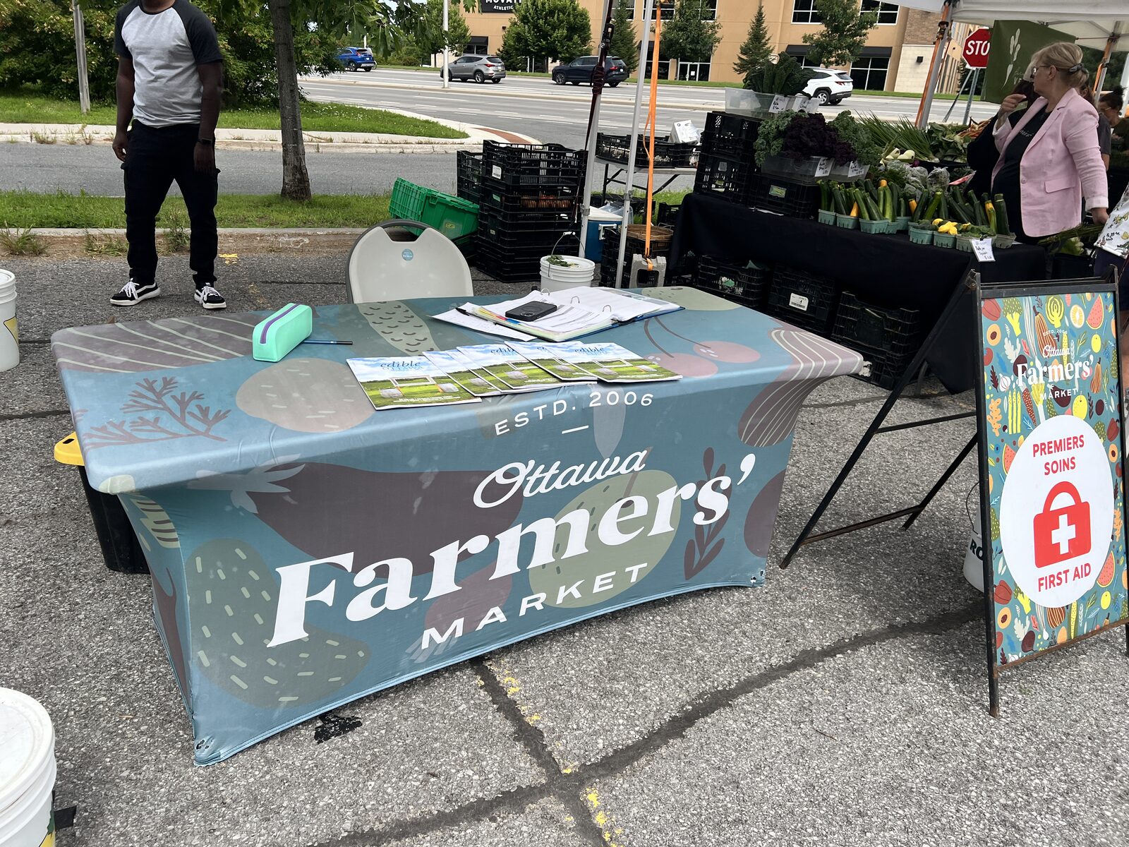 The Ottawa Farmers' Market information table, its cover reading Estd. 2006, with a produce stall behind it.