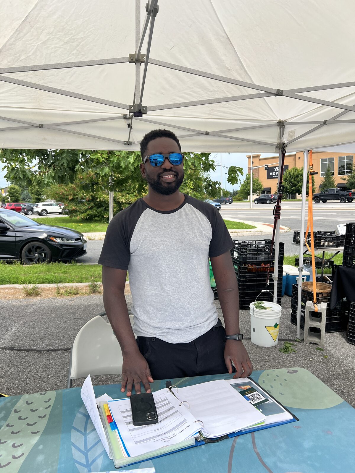 Eric Kyadondo standing behind the Ottawa Farmers' Market information table at the Barrhaven market, with the vendor binder open in front of him.