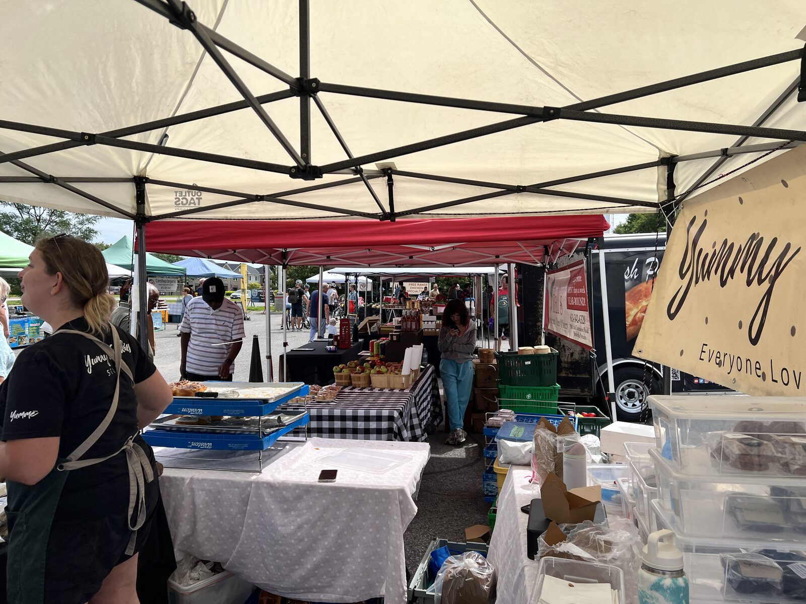 A bakery stall under a Yummy banner with trays of pastries, looking out across the market to a pie truck and crates of apples.