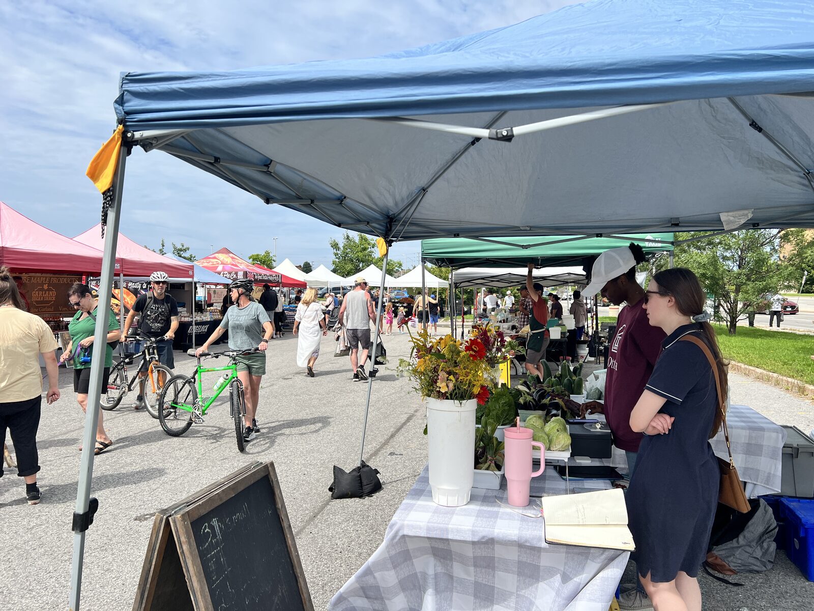 Shoppers and cyclists moving down the lane between tents, past a stall with buckets of cut flowers and crates of vegetables.
