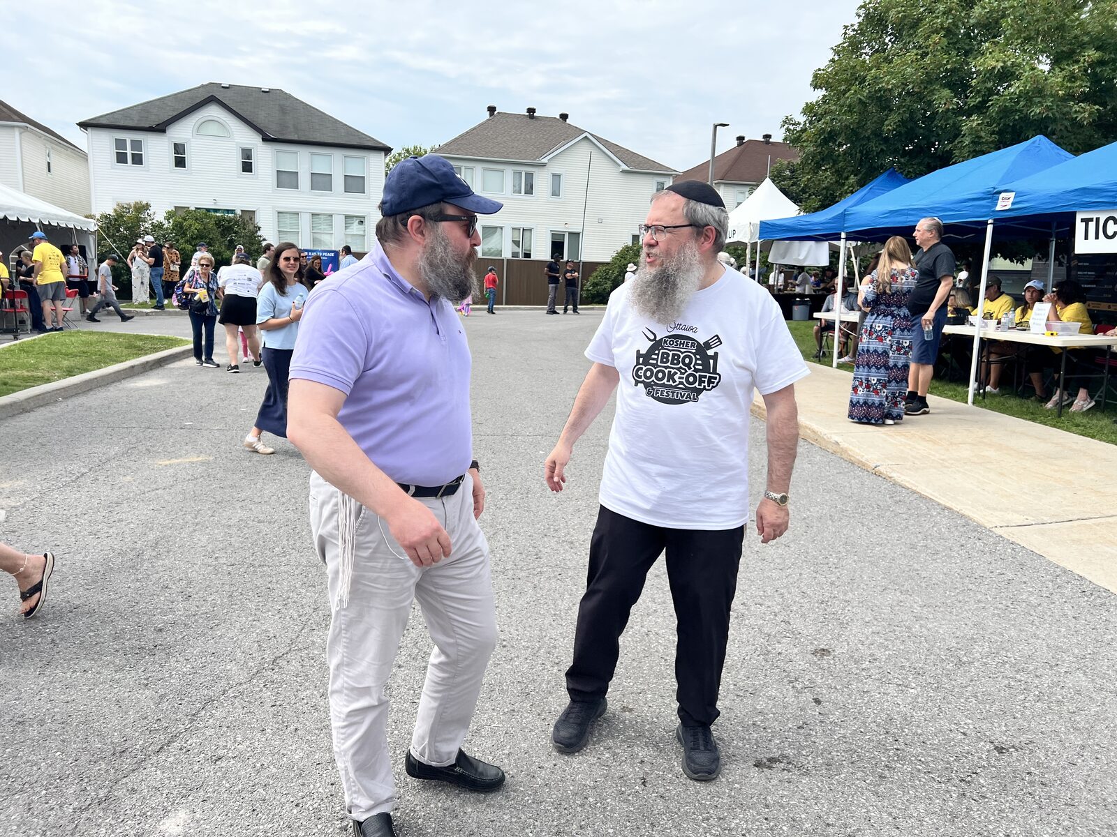 Two men in conversation on the closed street at the kosher BBQ cook-off, one wearing an Ottawa Kosher BBQ Cook-Off and Festival shirt.