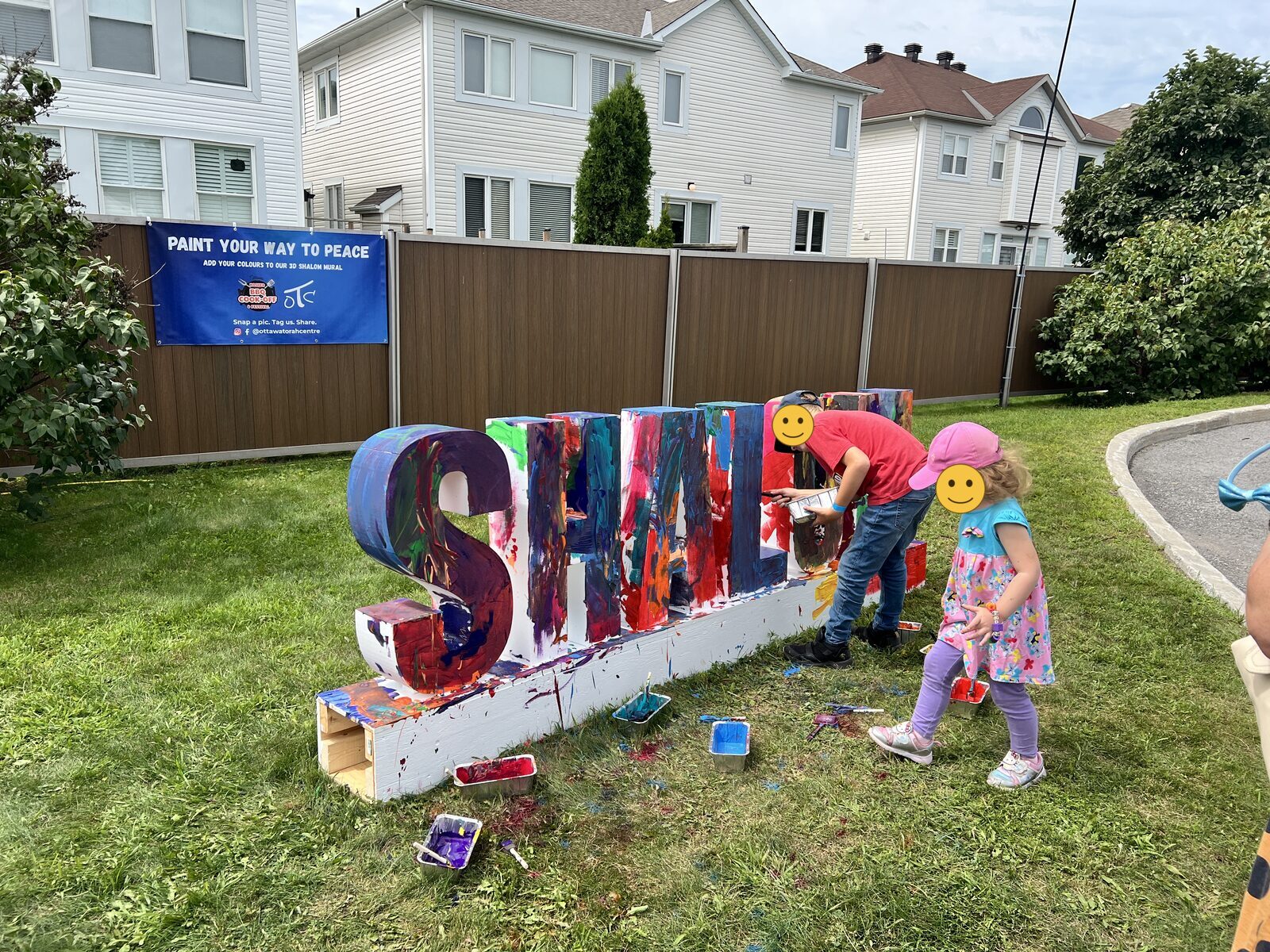 Two children painting large three-dimensional letters spelling Shalom on the grass, beneath a banner reading Paint Your Way to Peace.
