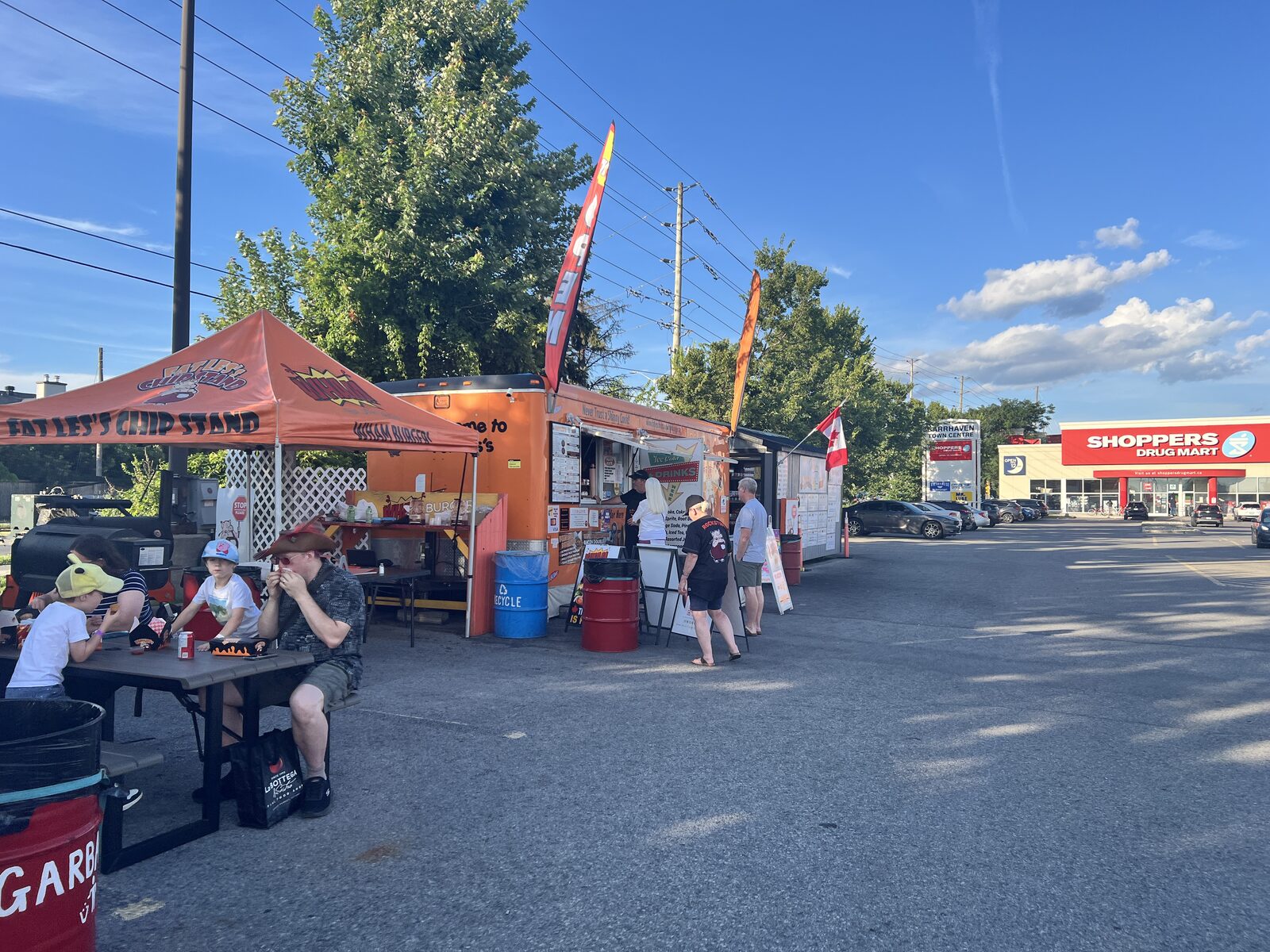 Customers lined up at the window of the orange Fat Les's Chip Stand trailer on Strandherd Drive, with picnic tables under an orange canopy and Shoppers Drug Mart across the lot.