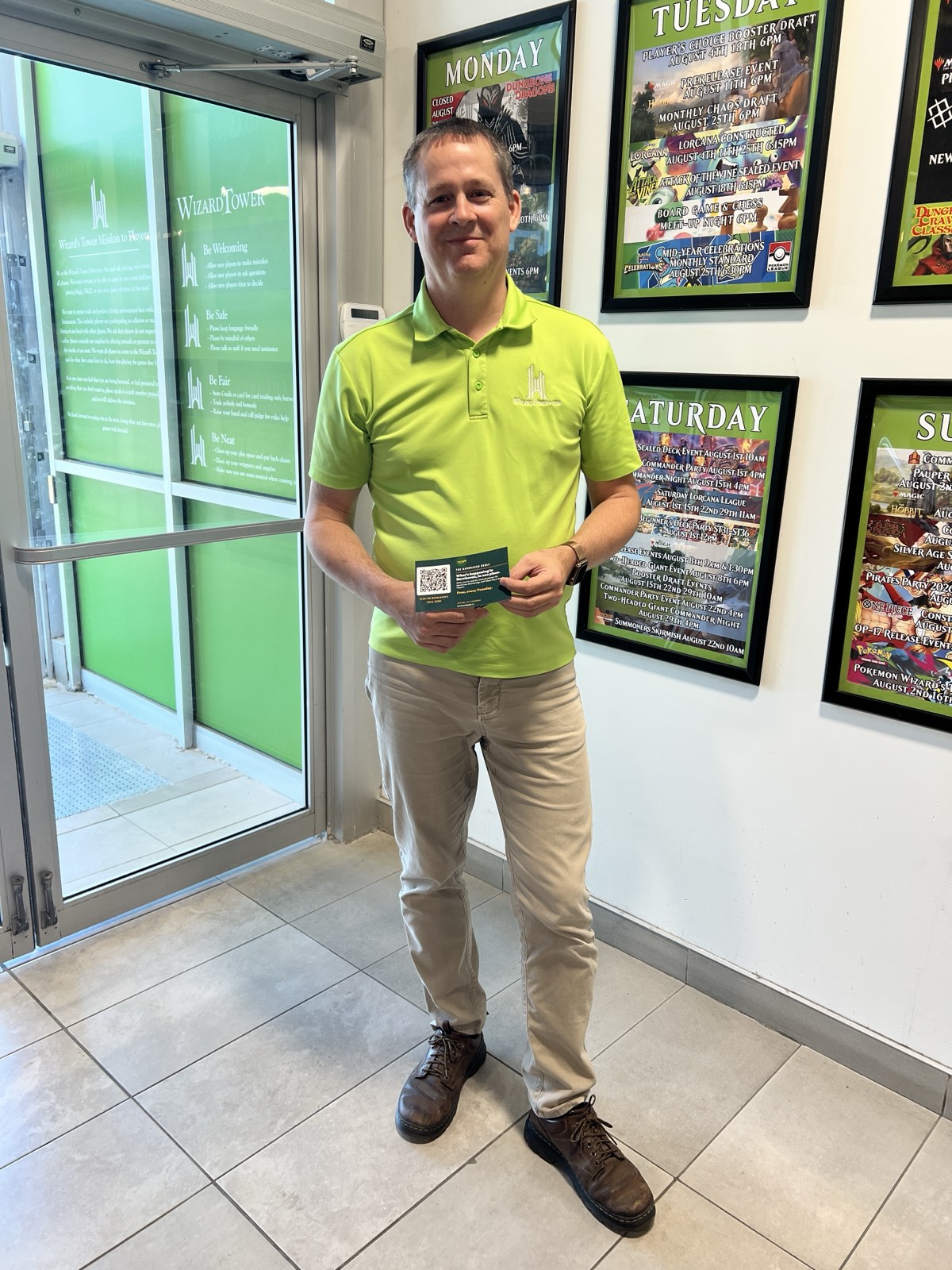 Dave, owner of Wizard's Tower, stands inside the Marketplace Avenue shop holding a Barrhaven Bugle card in front of the store's weekly events schedule.
